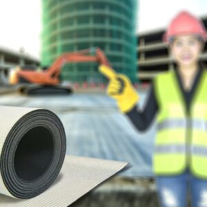 A close-up image of non-woven geotextile fabric being laid down during road construction. The fabric is a light brown color and is being unrolled across a prepared gravel base. This fabric will act as a separator and reinforcement layer, improving drainage and increasing the road's stability.